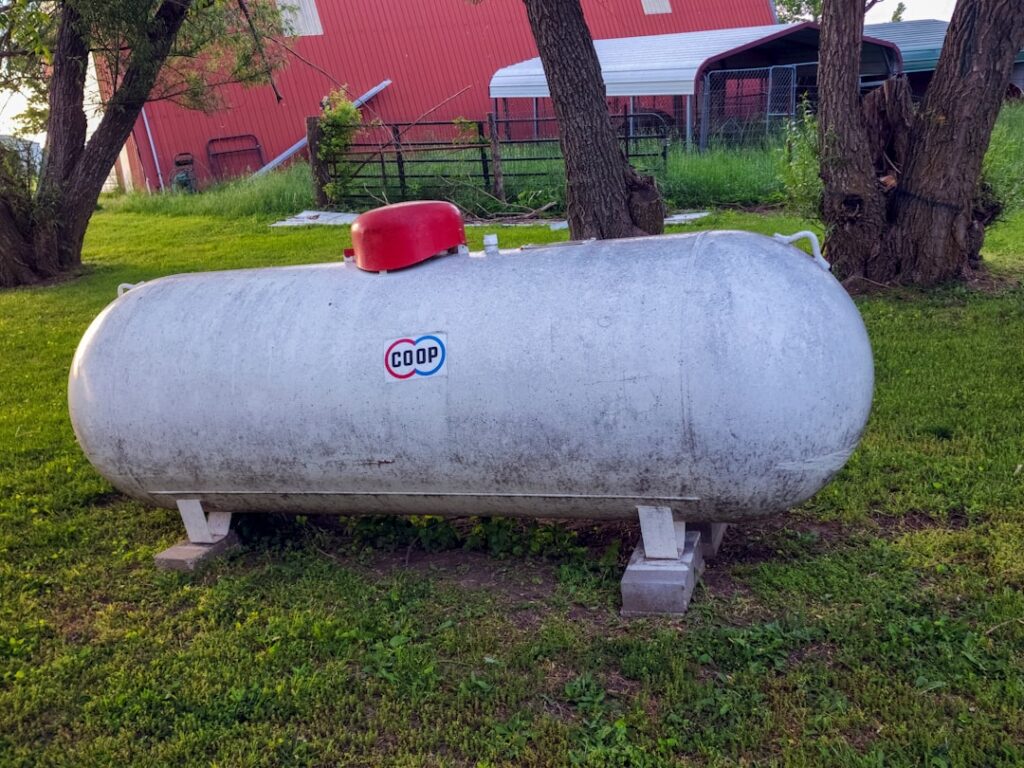 A backyard propane tank and utility gas meter next to a single-family home in a leafy residential neighborhood.